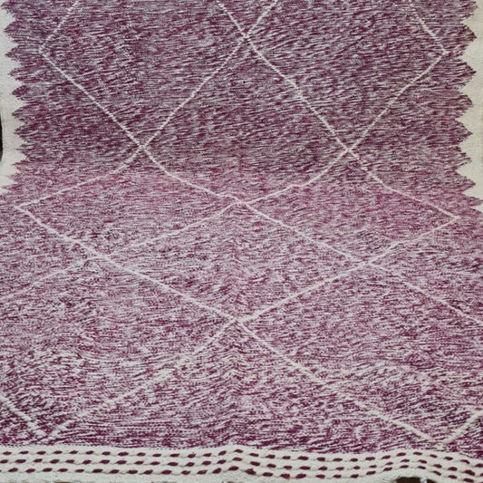 A rectangular woven rug with a dusty purple and cream geometric diamond pattern, featuring a thick cream border and fringe on the ends, displayed on a tiled floor.