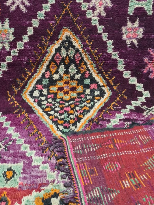 A traditional Moroccan-style wool rug with a deep purple field, featuring intricate geometric diamond medallions and floral motifs in orange, pink, yellow, green, and white. The rug is draped to show its detailed pattern and texture, with one corner folded over to reveal the reverse side.
