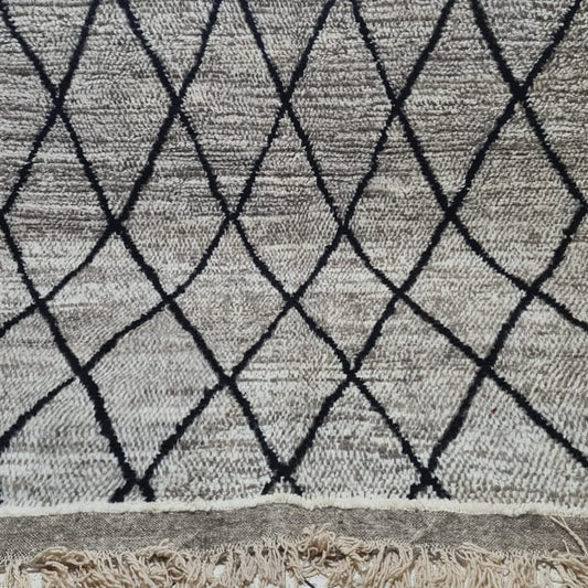 A rectangular woven throw blanket with a black diamond lattice pattern on a textured beige and off-white background, featuring fringed edges, displayed on a stand in an indoor setting.