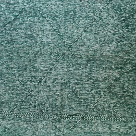 A large green woven rug with white fringe and a textured, heathered pattern, spread out on a tiled floor in front of a window.