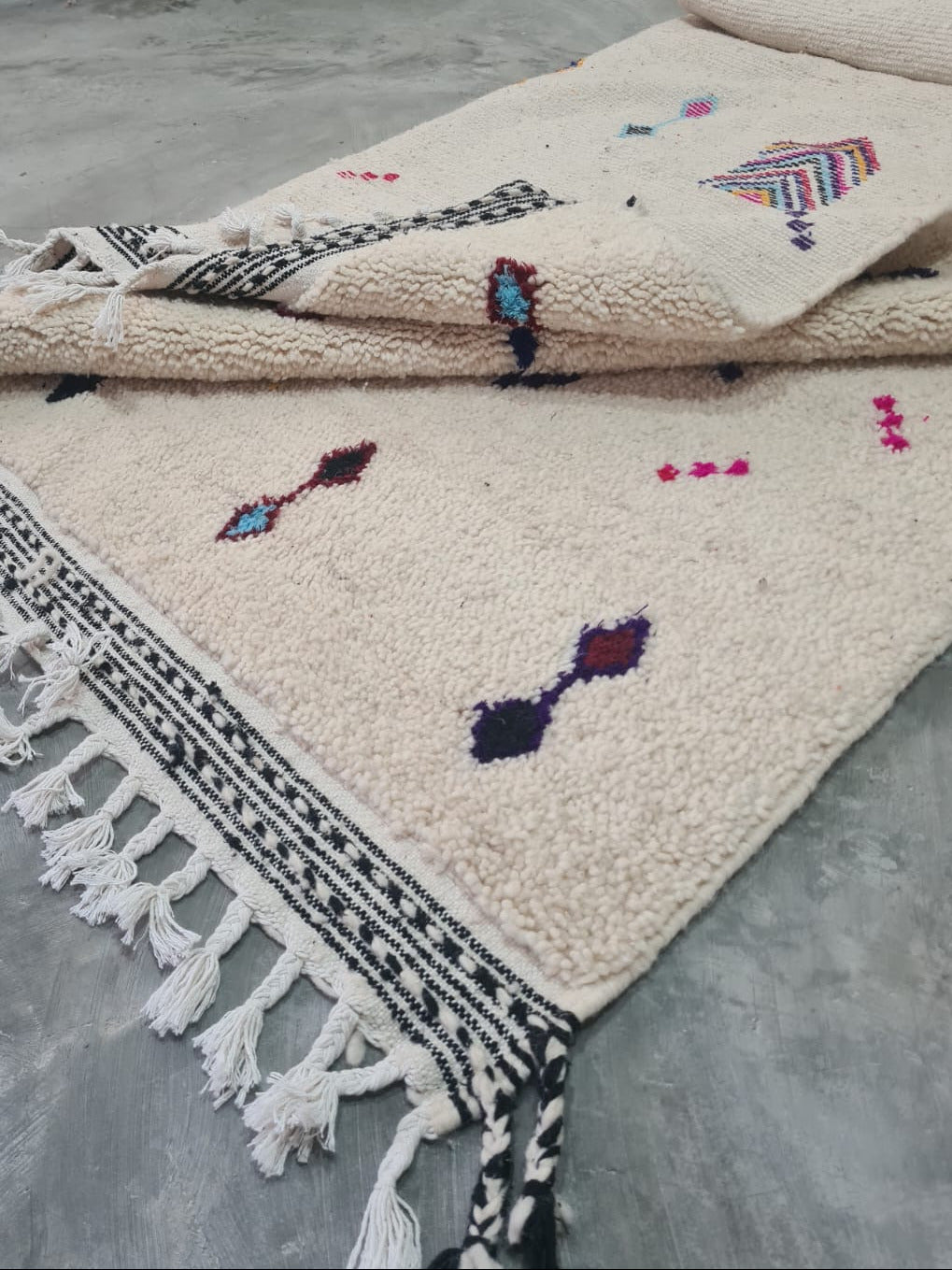 A cream-colored Moroccan-style wool rug with black geometric borders and multicolored embroidered motifs, laid out on a gray concrete floor.