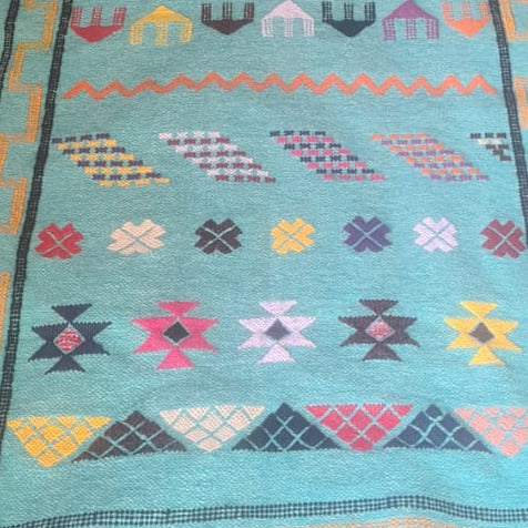 A turquoise blue woven runner rug with colorful geometric patterns and fringe, laid out on a textured brown and black patterned area rug in a home setting.