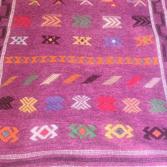 A purple woven runner rug with colorful geometric patterns, laid out on a patterned gray carpet in a home setting.