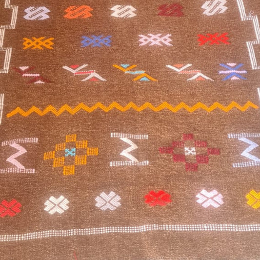 A long brown woven runner rug with colorful geometric patterns, laid out on a larger textured gray and black patterned area rug inside a home setting.