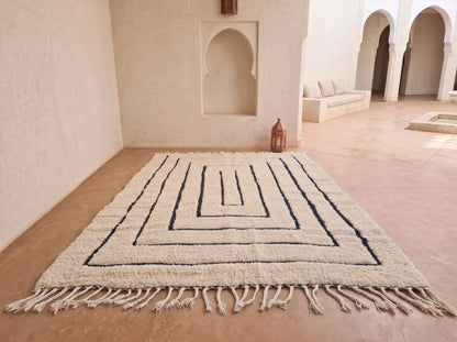 A large rectangular woven rug with a cream base and dark navy geometric maze-like border pattern, laid out on a terracotta tiled floor in a Moroccan-style room with arched alcoves and a bench in the background.