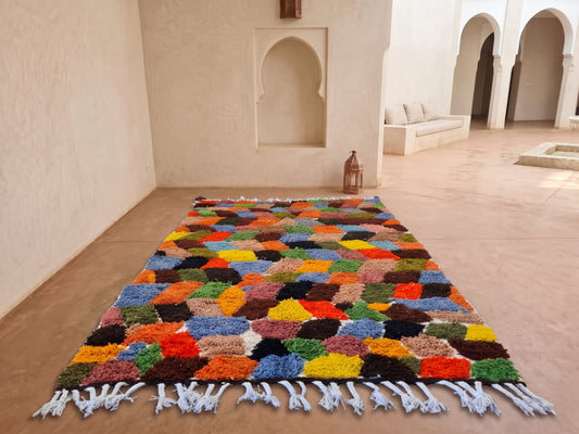 A colorful Moroccan shag rug with white fringe, displayed on a terracotta floor in a traditional Moroccan courtyard with arched alcoves.