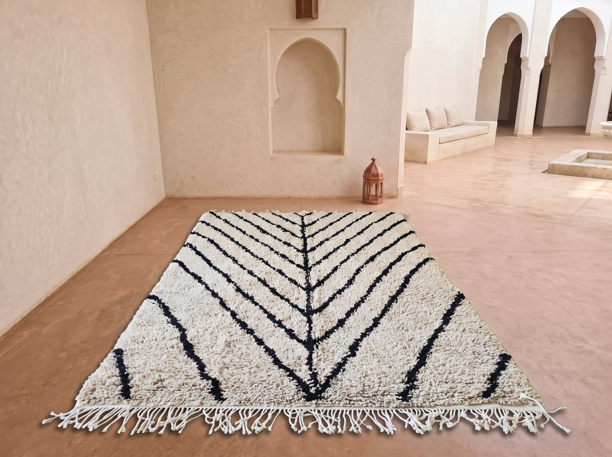 A large rectangular area rug with a cream and black chevron pattern and fringe, displayed on a terracotta floor in a Moroccan-style courtyard.