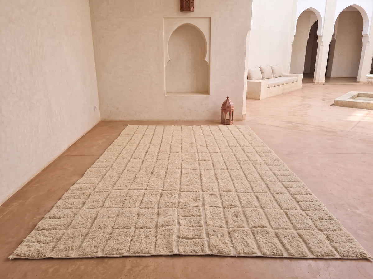 A large rectangular beige area rug with a raised grid pattern, displayed on a terracotta-colored tiled floor in a minimalist Moroccan-style room with cream walls and arches.