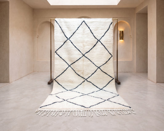 A large off-white Moroccan-style rug with a black diamond lattice pattern, displayed on a wooden stand in a minimalist beige room.
