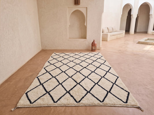 A large rectangular area rug with a cream base and a black geometric diamond pattern, displayed on a terracotta-colored floor in a Moroccan-style courtyard with arched alcoves and a small lantern.