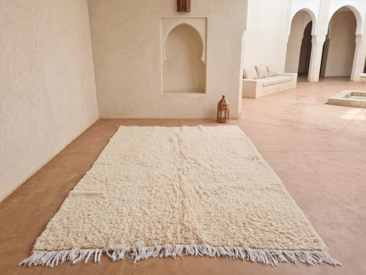 A large cream-colored shag rug with fringe trim laid out on a terracotta tiled floor in a Moroccan-style courtyard with beige walls and arches.
