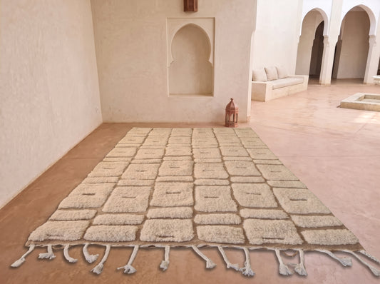 A large rectangular area rug with a raised grid pattern and fringe tassels, laid out on a terracotta tiled floor in a Moroccan-style courtyard with white stucco walls and arches.