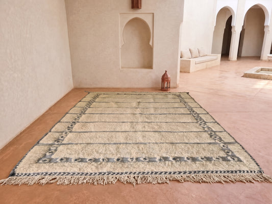 A rectangular woven rug with fringe and a geometric border pattern, displayed on a terracotta tiled floor in a Moroccan-style courtyard with beige walls and arches.