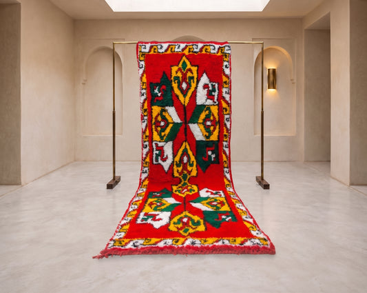 A large red woven rug with a bold geometric and floral pattern in yellow, green, white, and black, hanging from a metal stand in a minimalist beige gallery space.