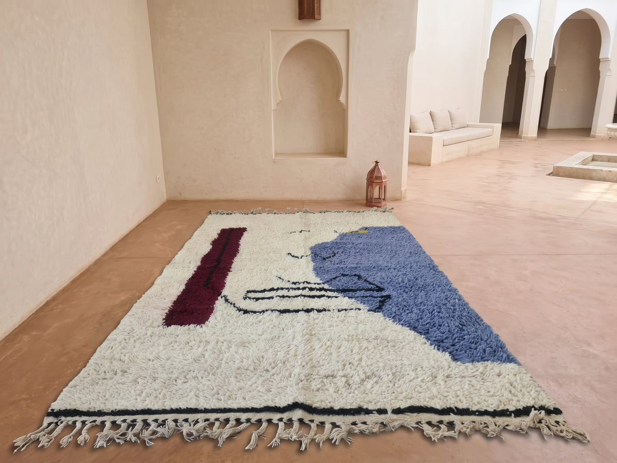 A large Moroccan-style shag rug with a cream base, a blue abstract shape, a dark red stripe, and black lines, displayed on a terracotta floor in a minimalist room with arched alcoves.