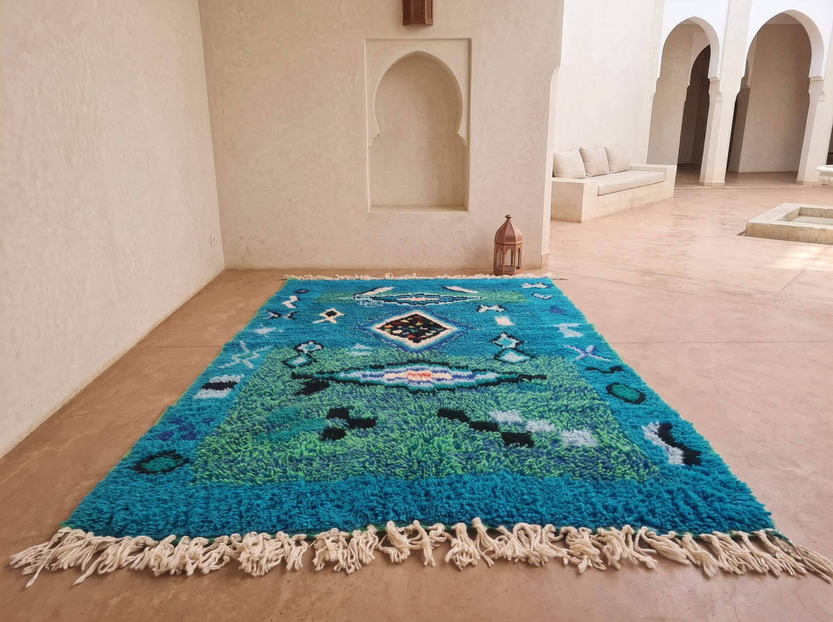 A rectangular shag rug with cream fringe, featuring a blue and green Moroccan-style geometric pattern, displayed on a terracotta tiled floor in a minimalist, warm-toned room with arched alcoves.