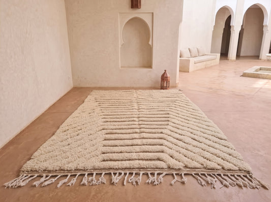 A large cream-colored Moroccan-style rug with a textured chevron pattern and fringe tassels, displayed on a terracotta-colored floor in a minimalist room with arched alcoves.