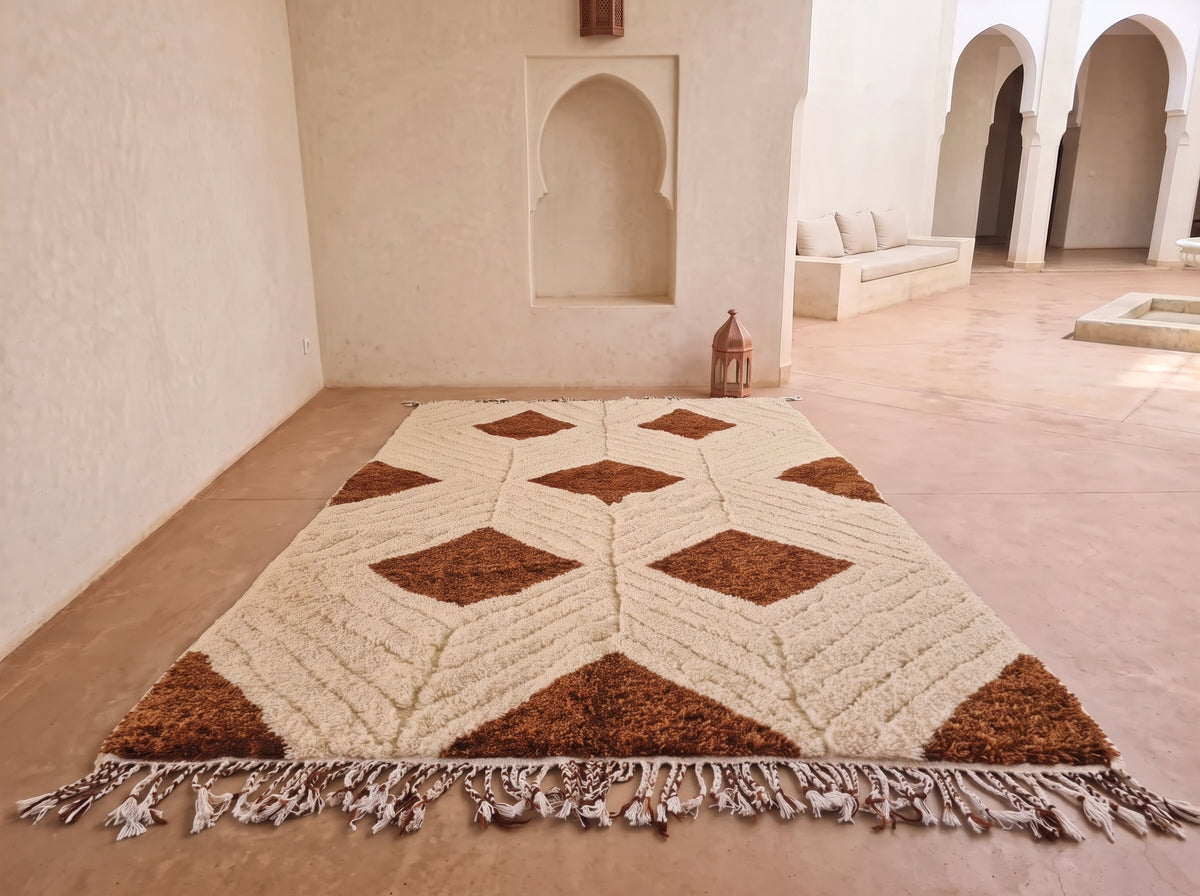 A rectangular area rug with a geometric diamond pattern in brown and cream, laid out on a terracotta tiled floor in a Moroccan-style courtyard with arched alcoves and a white bench in the background.