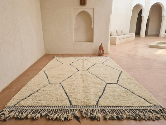 A large rectangular Moroccan-style rug with a cream and beige geometric pattern and fringed edges, displayed on a terracotta floor in a minimalist, sunlit room with arched alcoves.