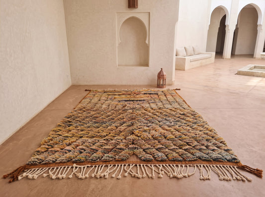 A large Moroccan-style woven area rug with a textured geometric pattern in earthy tones of brown, beige, and muted blue, laid out on a terracotta tiled floor in a minimalist courtyard with white stucco walls and arches.