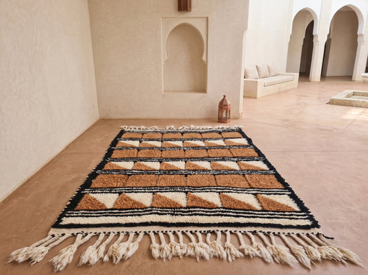 A rectangular Moroccan-style woven rug with cream, tan, and black geometric patterns and fringe tassels, displayed on a terracotta floor in a minimalist, sunlit courtyard with arches.