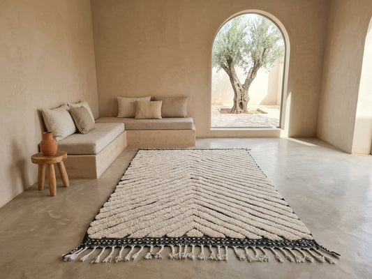 A large rectangular area rug with a textured chevron pattern in cream and black, featuring fringe on one end, styled in a minimalist, warm-toned modern room with a built-in bench and an arched window view of an olive tree.