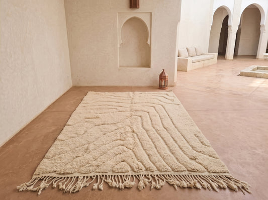 A large cream-colored Moroccan-style rug with a textured wavy pattern and tasseled fringe, displayed on a terracotta-colored floor in a minimalist, sunlit courtyard with white stucco walls and arches.