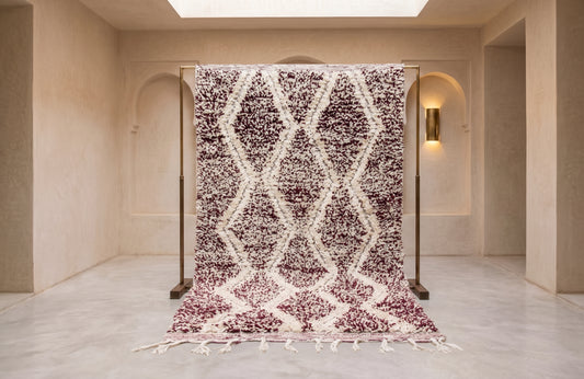 A large Moroccan-style area rug with a textured geometric diamond pattern in dark plum and off-white, displayed hanging from a wooden frame in a minimalist beige hallway.