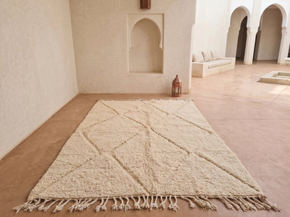 A large cream-colored Moroccan-style wool rug with a geometric diamond pattern and fringe tassels, laid out on a terracotta-colored floor in a minimalist, sunlit courtyard with arched walls.