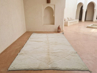 A large off-white Moroccan rug with a diamond-quilted pattern laid out on a terracotta floor in a minimalist, sunlit room with arched alcoves.