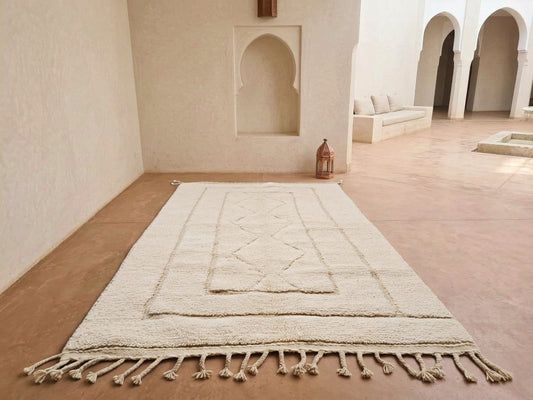 A large cream-colored Moroccan-style area rug with geometric woven patterns and tasseled fringe, displayed on a terracotta-colored floor in a minimalist, sunlit room with arched alcoves.