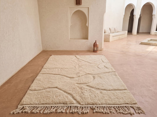 A large cream-colored Moroccan-style area rug with a textured raised pattern and fringe, displayed on a terracotta-colored floor in a minimalist, sunlit courtyard with arched walls.