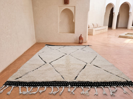 A large rectangular Moroccan-style rug with a cream shaggy texture, black geometric diamond lines, and a black-and-white dotted fringe, displayed on a terracotta tiled floor in a minimalist courtyard with beige walls and arches.