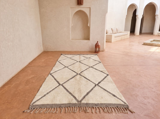 A rectangular beige rug with a dark charcoal geometric diamond pattern and tasseled fringe, displayed on a terracotta-colored tiled floor in a Moroccan-style courtyard.