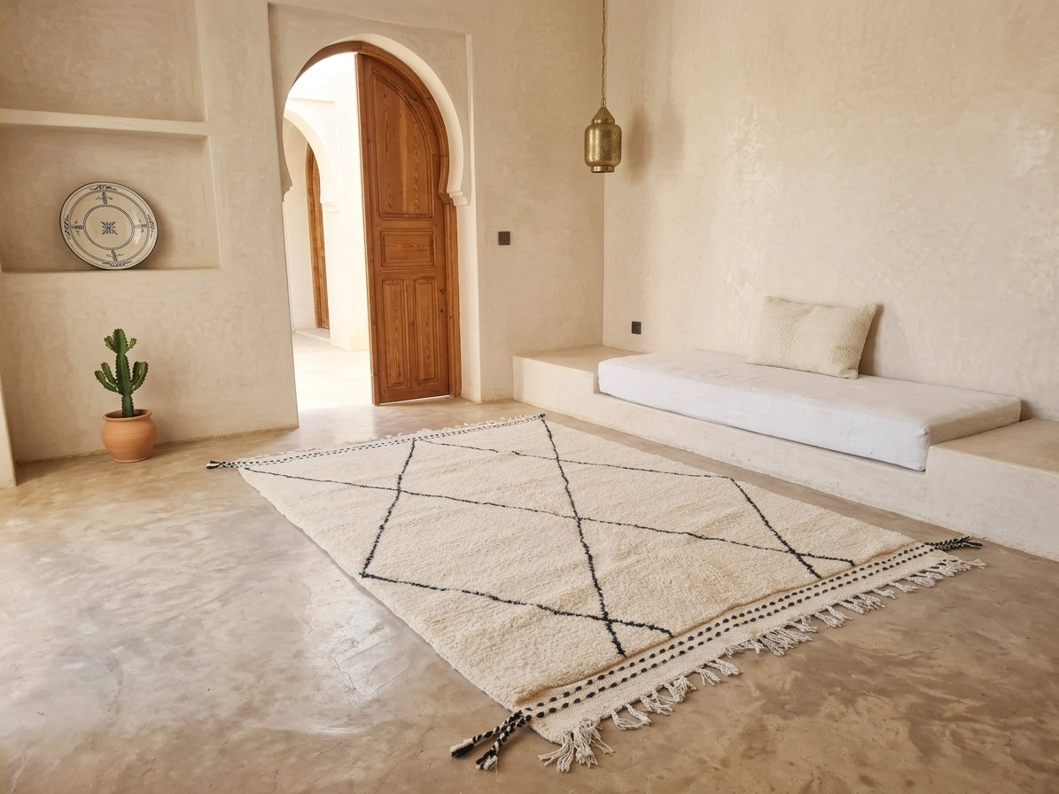 A cream-colored Moroccan-style area rug with black geometric lines and fringe, displayed on a stone floor in a minimalist, warm-toned room with a built-in bench and wooden door.