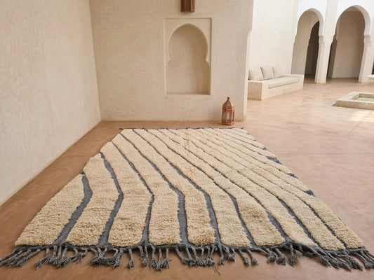 A large rectangular area rug with a textured, shaggy cream and dark gray chevron pattern and tasseled fringe, displayed on a terracotta tiled floor in a Moroccan-style courtyard.