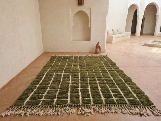 A rectangular green and beige woven rug with fringe, displayed on a terracotta tiled floor in a Moroccan-style room with arched alcoves and a bench.