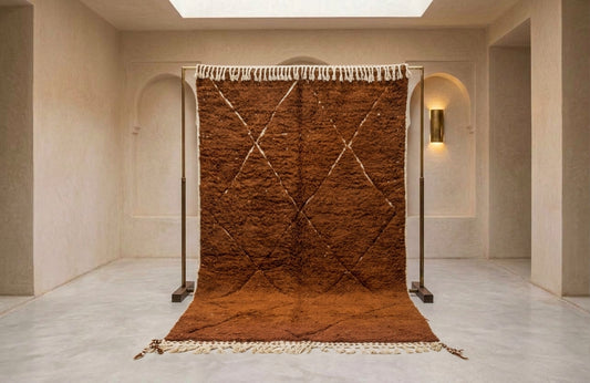 A large brown Moroccan-style rug with cream fringe and a diamond pattern, displayed on a black metal stand in a minimalist beige room.