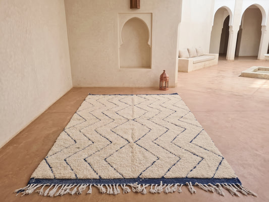 A rectangular Moroccan-style wool rug with a cream and beige geometric zigzag pattern and a dark blue border with fringe, displayed on a terracotta floor in a minimalist, sunlit courtyard.