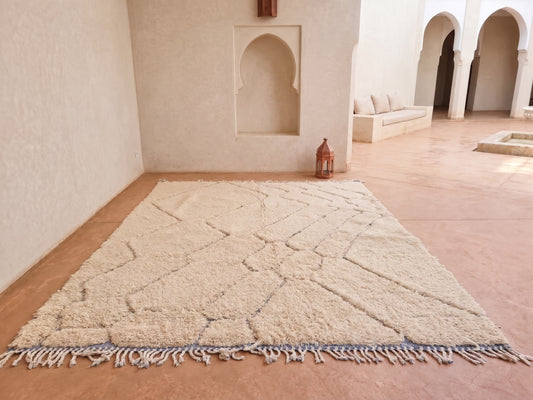 A large off-white Moroccan-style shag rug with a geometric pattern and fringed edge, laid out on a terracotta tiled floor in a minimalist courtyard with cream walls and arches.