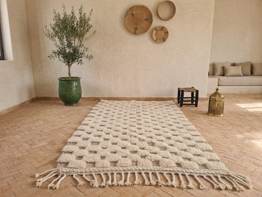 A cream-colored textured woven area rug with fringe, laid out on a herringbone wood floor in a warm, boho-style room with a potted olive tree and wall decor.