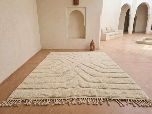 A large cream-colored Moroccan-style area rug with a textured geometric pattern and fringe, displayed on a terracotta floor in a minimalist, sunlit room with arched alcoves.