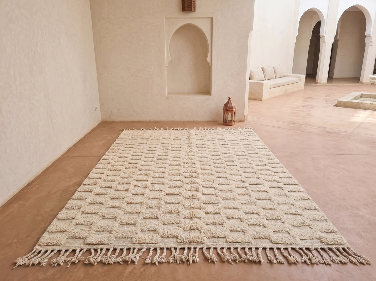 A large cream-colored textured area rug with fringe, displayed on a terracotta-colored floor in a minimalist Moroccan-style room with arched alcoves.