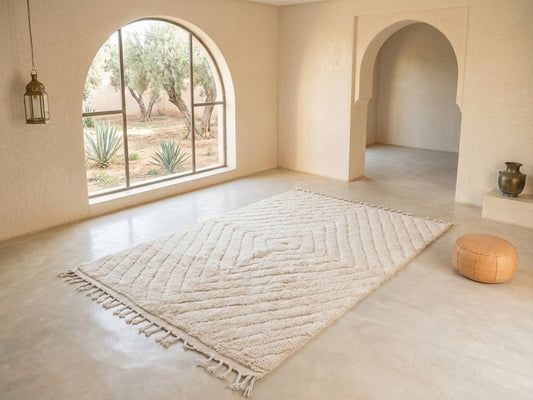 A large cream-colored textured area rug with fringe tassels and a geometric diamond pattern, displayed in a minimalist, warm-toned room with arched windows and walls.