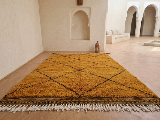 A large rectangular shag rug in mustard yellow with a black diamond pattern and white fringe, displayed on a beige stone floor in a minimalist Moroccan-style room.