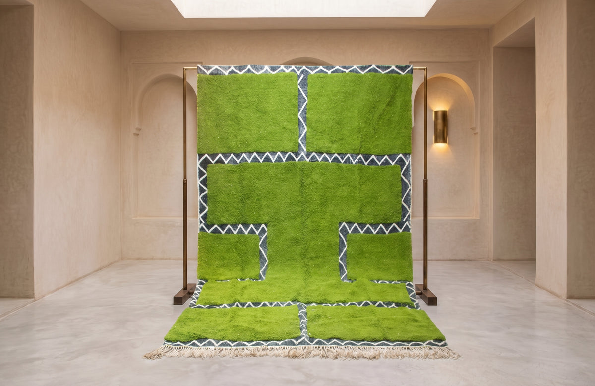 A bright green Moroccan-style rug with geometric patterns and white zigzag borders, displayed on a wooden stand in a minimalist beige room.