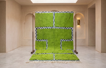 A bright green Moroccan-style rug with geometric patterns and white zigzag borders, displayed on a wooden stand in a minimalist beige room.