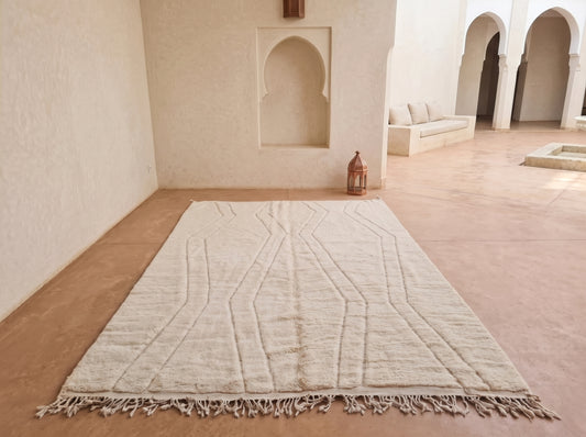 A large off-white Moroccan wool area rug with geometric woven patterns and fringed edges, displayed on a terracotta tiled floor in a minimalist, sunlit courtyard with beige walls and arches.