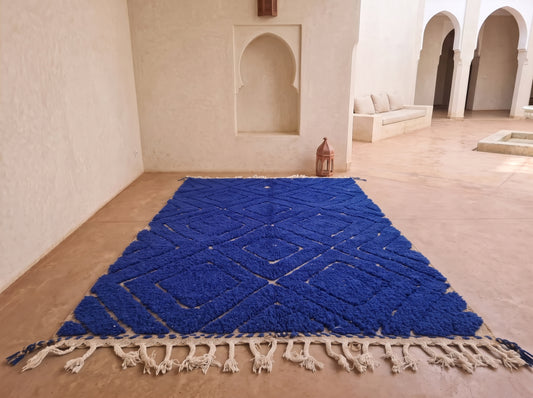 A vibrant royal blue Moroccan-style rug with a textured geometric pattern and cream-colored fringe, displayed on a terracotta floor in a minimalist, sunlit courtyard with white stucco walls and arches.