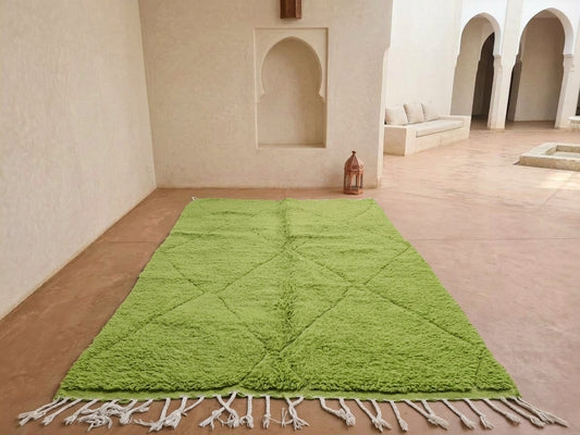 A bright green Moroccan-style shag rug with white fringe tassels, laid out on a terracotta tiled floor in a minimalist, sunlit courtyard with white stucco walls and arches.